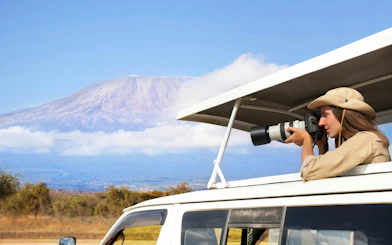 Woman photographing wildlife from safari vehicle with Mount Kilimanjaro in background, Kenya.