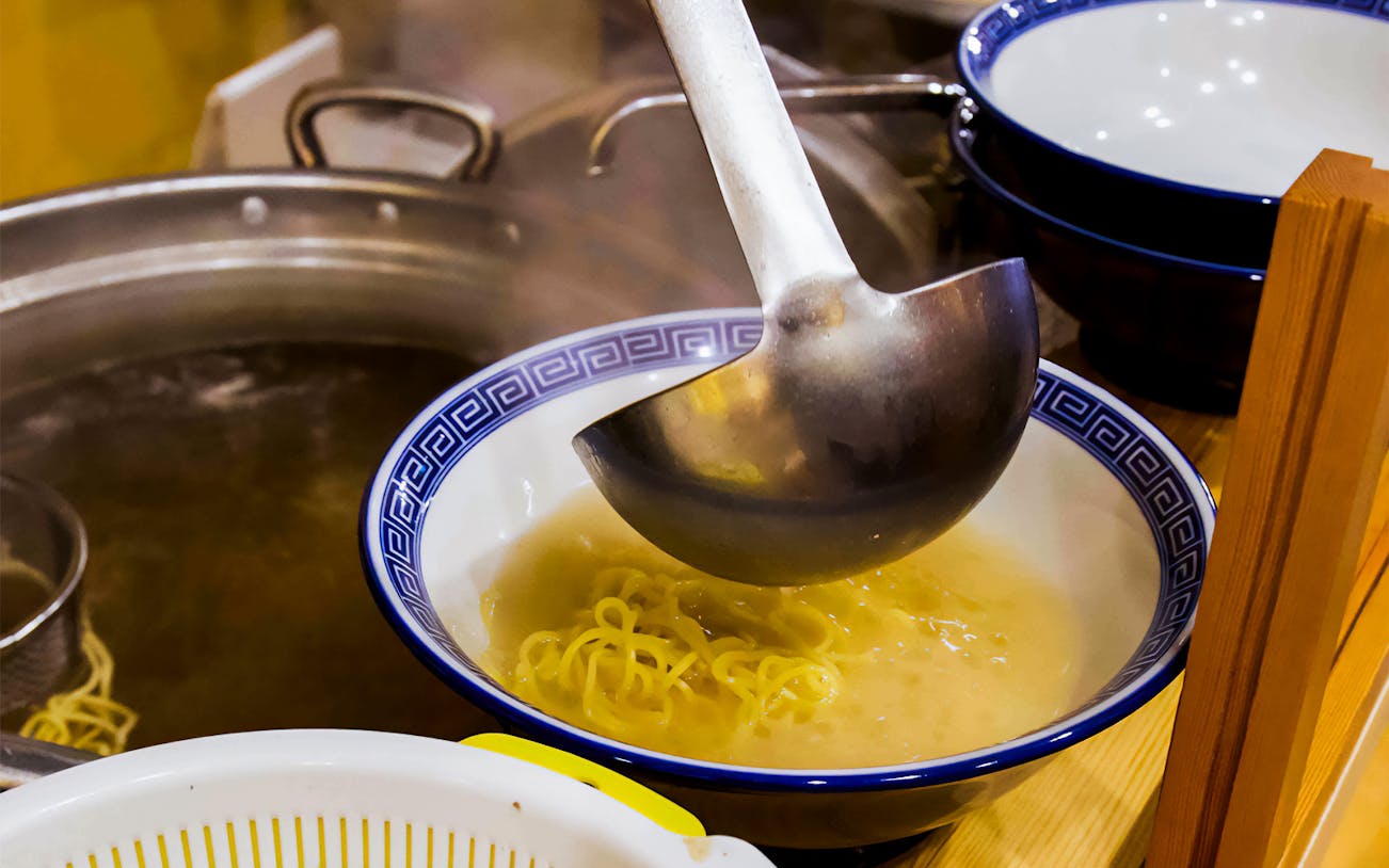 Ladle pouring broth over noodles in a ramen kitchen experience.