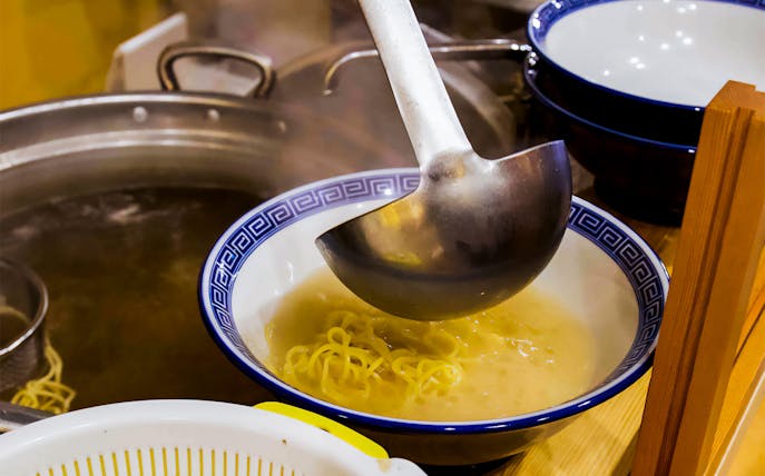 Ladle pouring broth over noodles in a ramen kitchen experience.