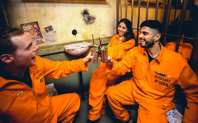 Guests in orange jumpsuits enjoying cocktails at Alcotraz prison-themed bar.