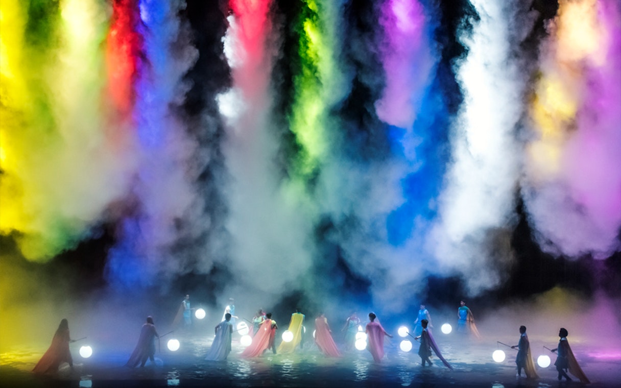 Performers with glowing spheres on a misty stage with colorful lights above.