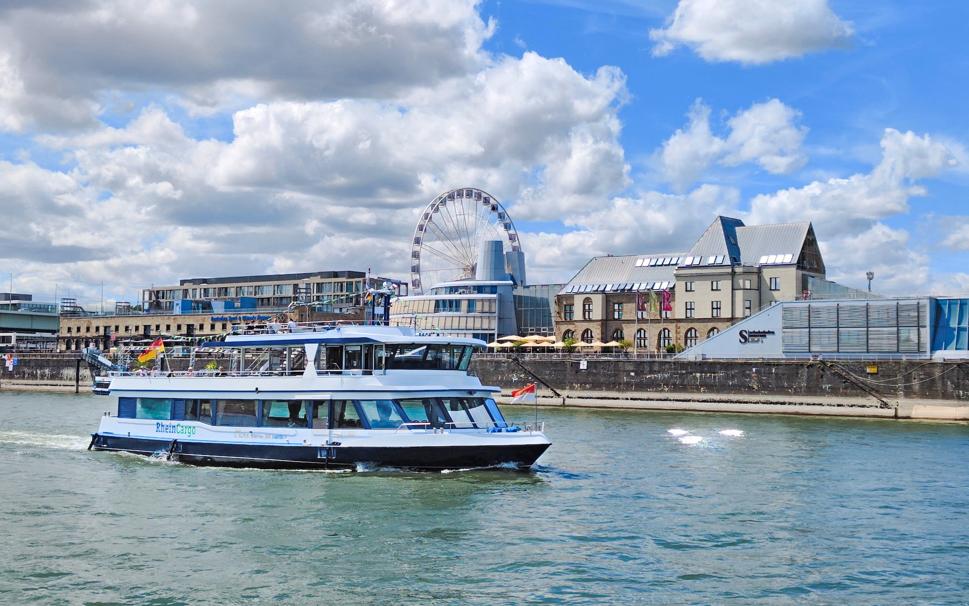 Cruise boat near Cologne Chocolate Museum with Ferris wheel in background.