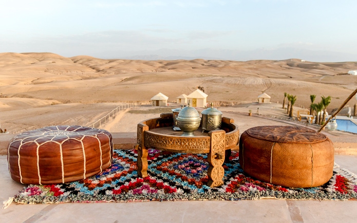 Tea setup on colorful rug in Agafay Desert, Marrakesh with poufs and desert tents in background.