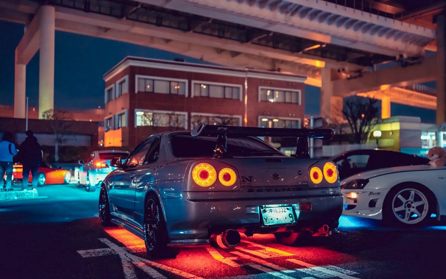 Tokyo Daikoku PA night car meeting with illuminated sports cars under highway.