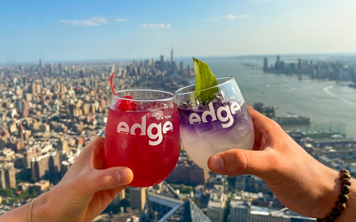 Two people toasting with drinks at Edge, New York City skyline in the background.