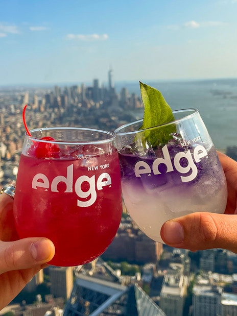 Two people toasting with drinks at Edge, New York City skyline in the background.