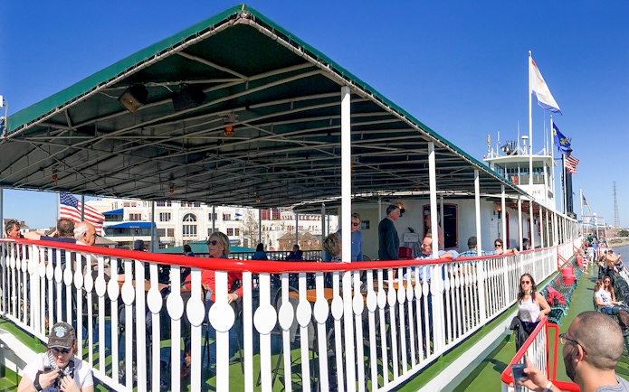 Steamboat Natchez deck with passengers, New Orleans skyline in background on Mississippi River.