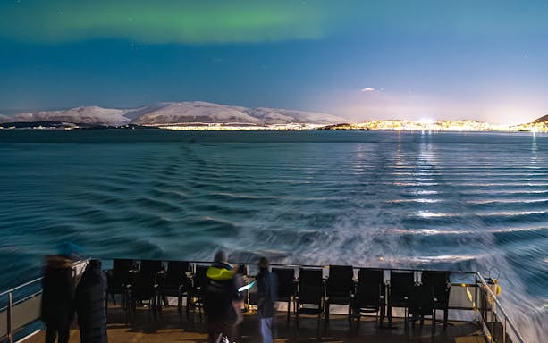 Cruise ship deck under Northern Lights with view of snowy mountains and city lights.