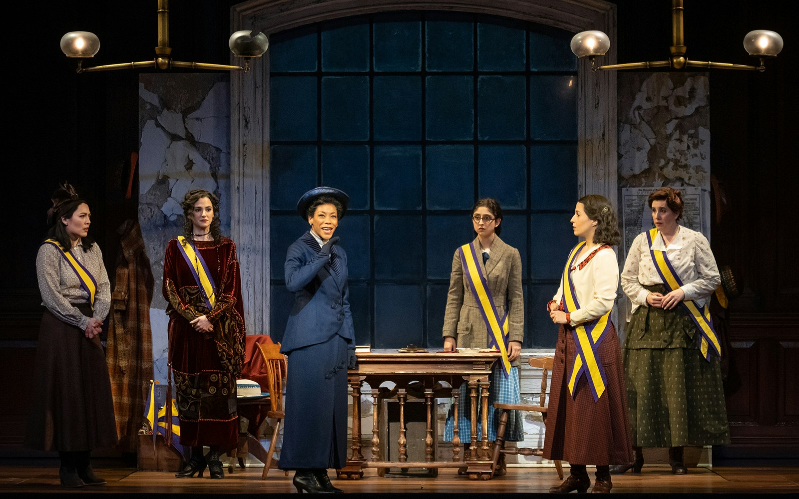 Women in period costumes with sashes on stage during a performance of "Suffs.