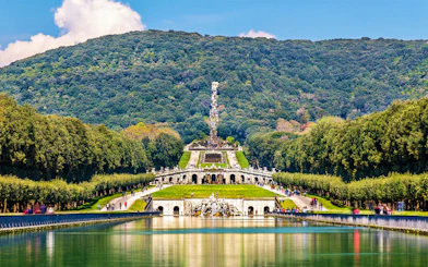Royal Palace of Caserta gardens with fountain and wooded hills in the background.