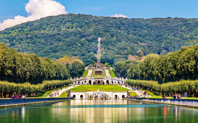 Royal Palace of Caserta gardens with fountain and wooded hills in the background.