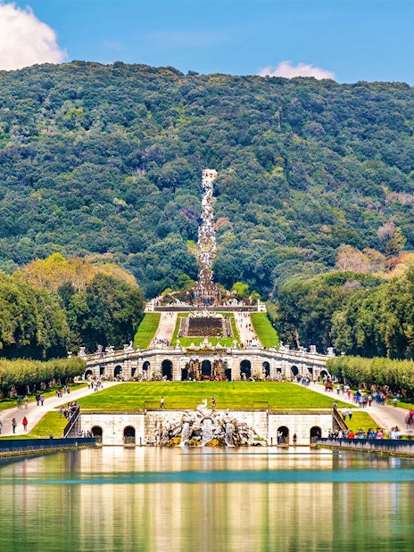 Royal Palace of Caserta gardens with fountain and wooded hills in the background.