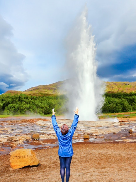 Woman raising arms near erupting Strokkur Geyser, Iceland.