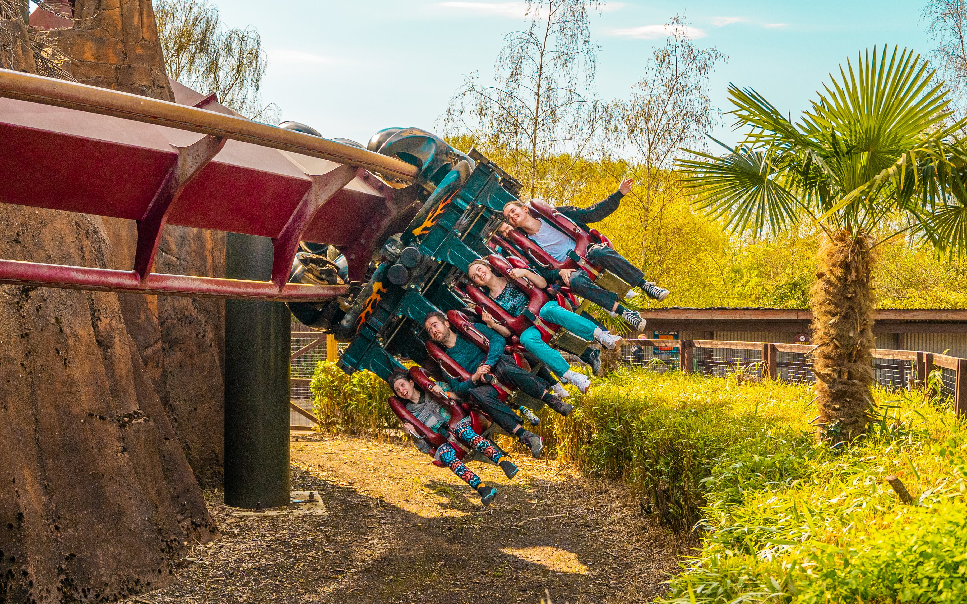 Roller coaster riders at Thorpe Park on an inverted track amidst greenery.