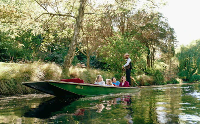 Punting on the Avon River in Christchurch with a guide and passengers surrounded by lush greenery.