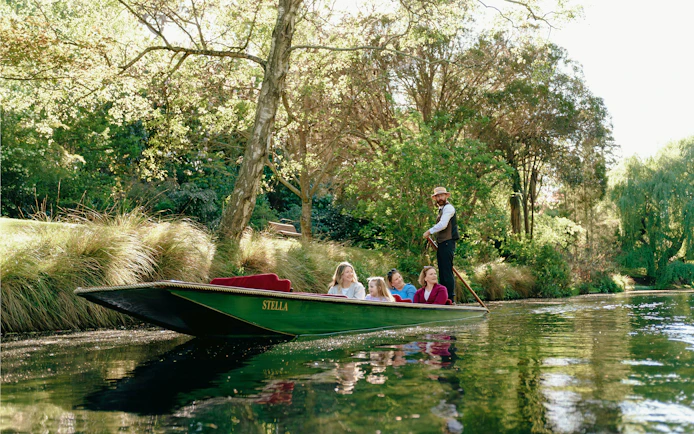 Punting on the Avon River in Christchurch with a guide and passengers surrounded by lush greenery.