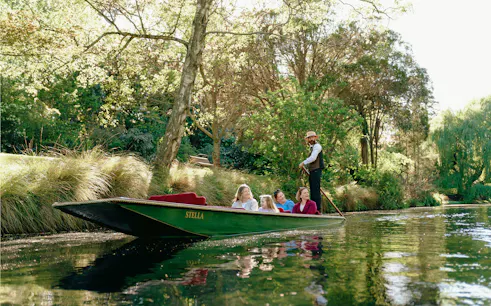 Punting on the Avon River in Christchurch with a guide and passengers surrounded by lush greenery.