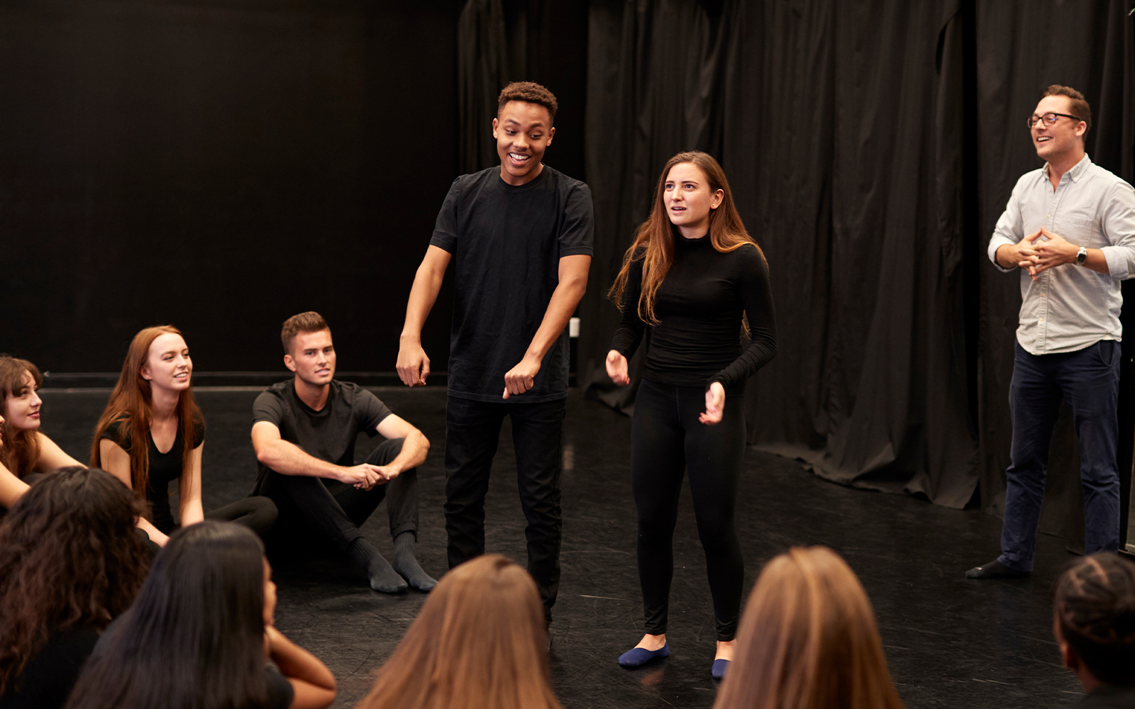 Theatre actors performing in front of a seated audience in a black box theater.