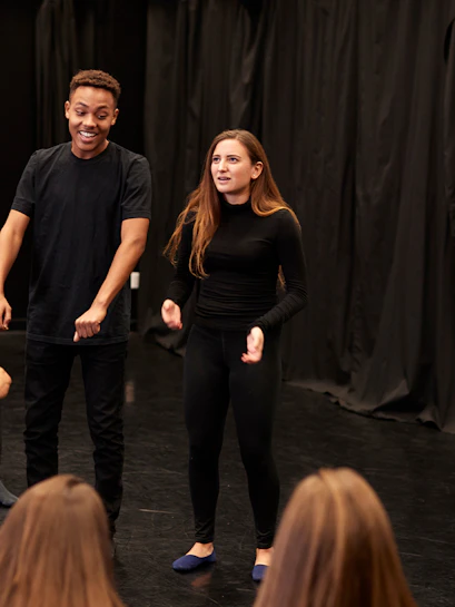 Theatre actors performing in front of a seated audience in a black box theater.