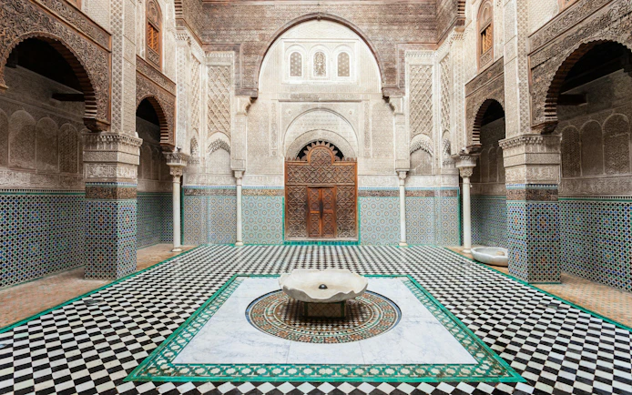 Al Attarine Madrasa courtyard with intricate tilework and carved wooden door in Fez, Morocco.