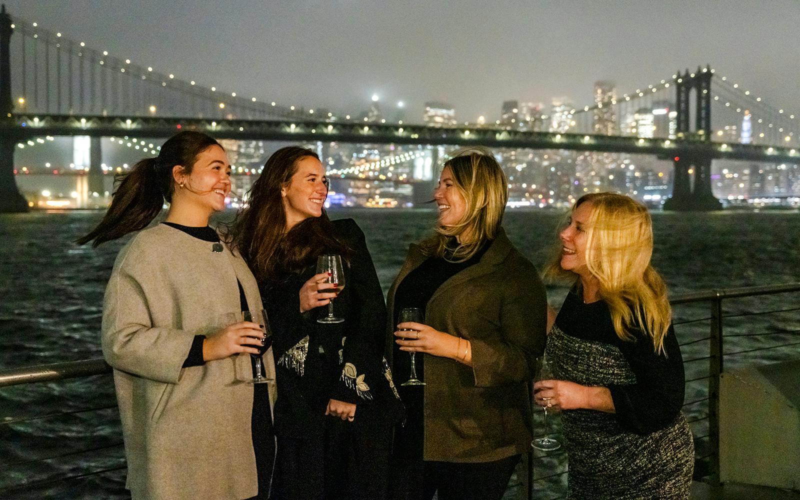 Women enjoying drinks on a New Jersey Signature Cruise with city skyline and bridge in the background.