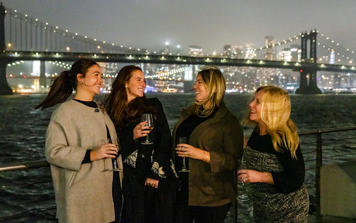 Women enjoying drinks on a New Jersey Signature Cruise with city skyline and bridge in the background.