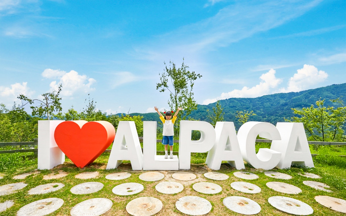 Girl standing on "I Love Alpaca" sign at Alpaca Island with mountains in background.
