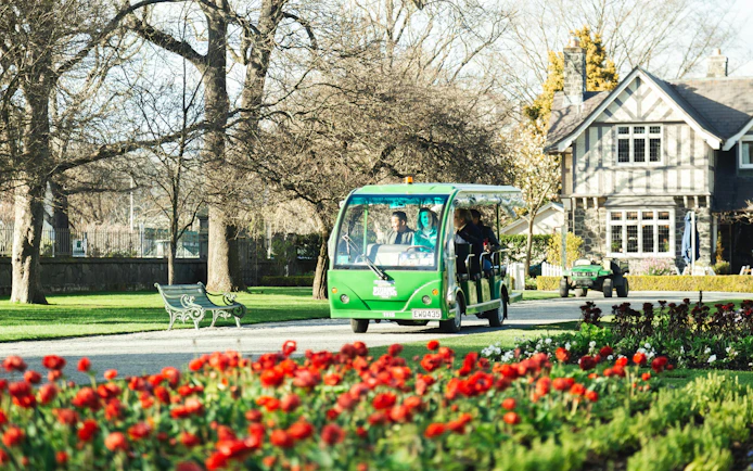 Tour tram in Christchurch Botanic Gardens with blooming flowers and historic building.