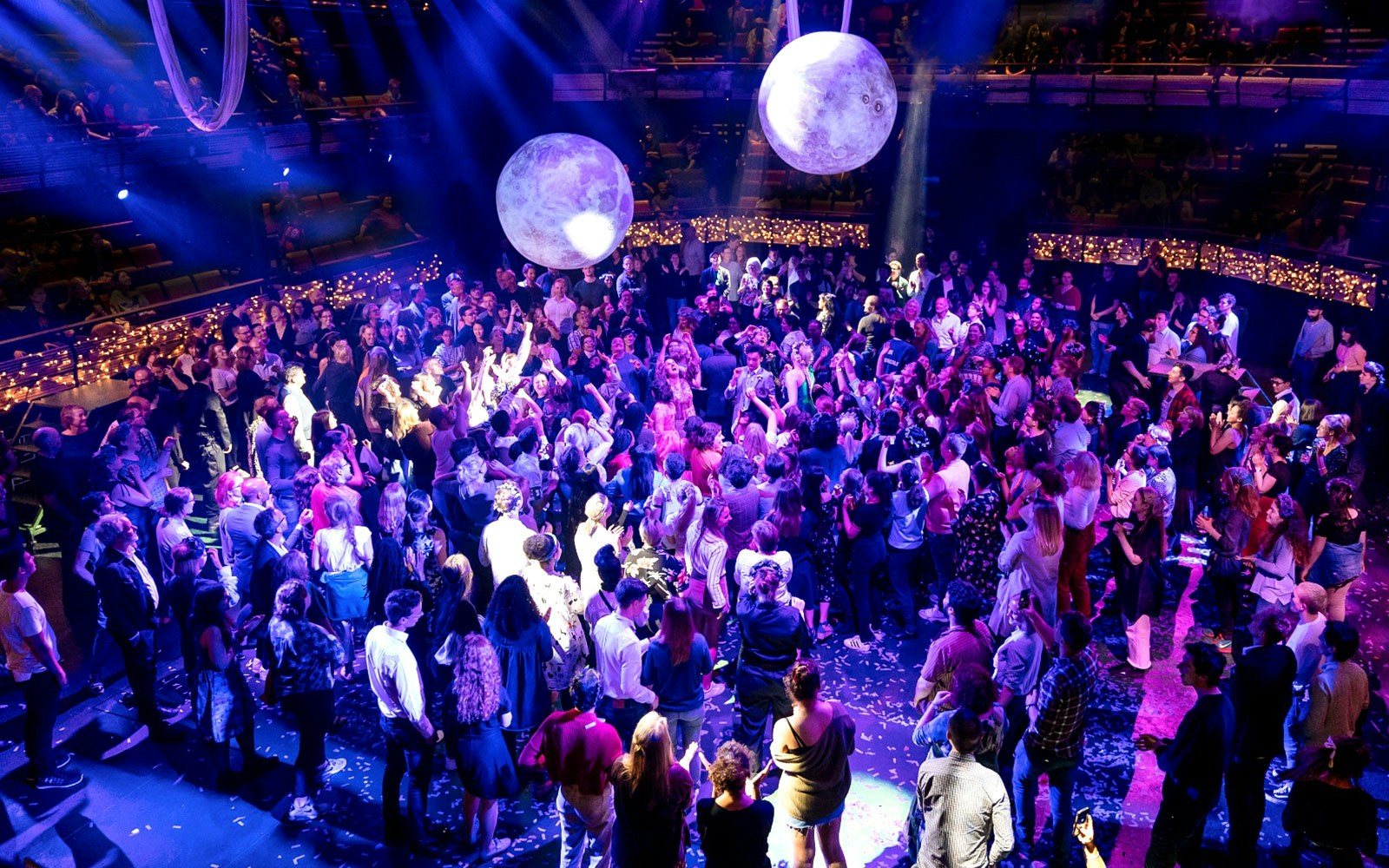 Crowd enjoying a performance of A Midsummer Night’s Dream with illuminated orbs above.