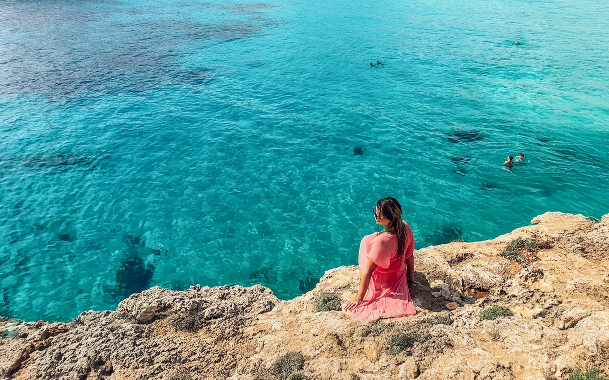 Tourist sitting on rocky shore overlooking Blue Lagoon, Malta.