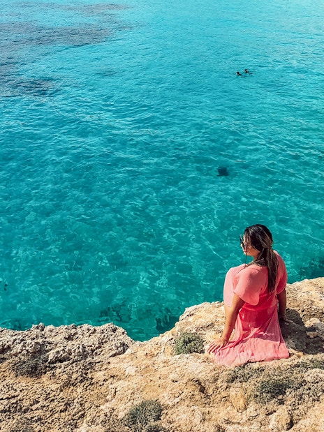 Tourist sitting on rocky shore overlooking Blue Lagoon, Malta.
