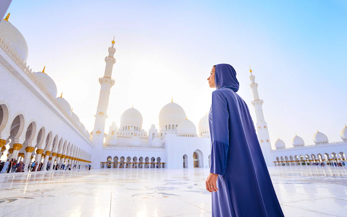 Woman in abaya at Sheikh Zayed Grand Mosque, Abu Dhabi, with domes and minarets in view.