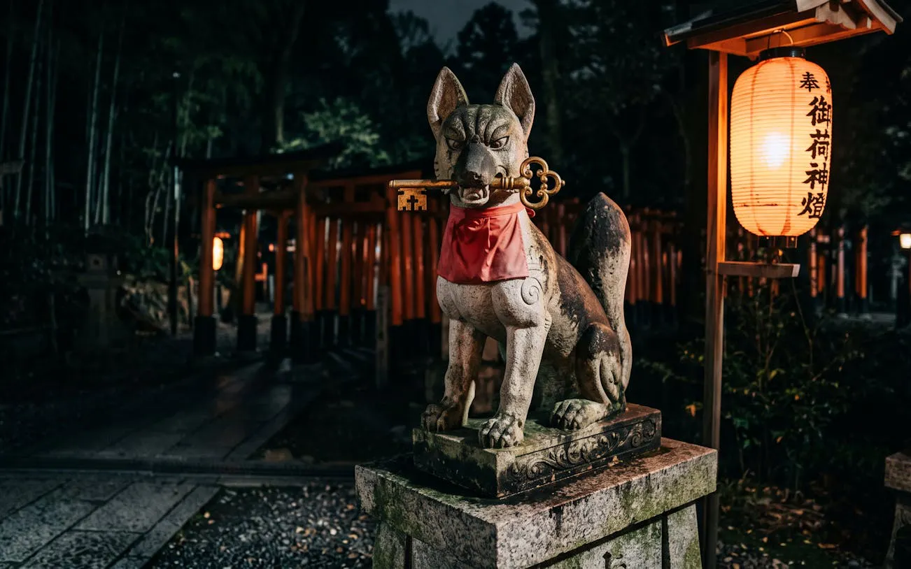 Stone fox statue with key at Fushimi Inari, Kyoto, illuminated by lanterns at night.