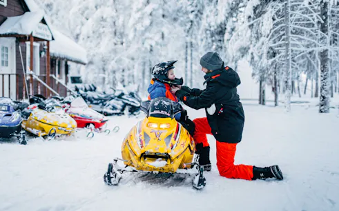 Man adjusting child's helmet on snowmobile at Arctic Circle Snowmobile Park, Finland.