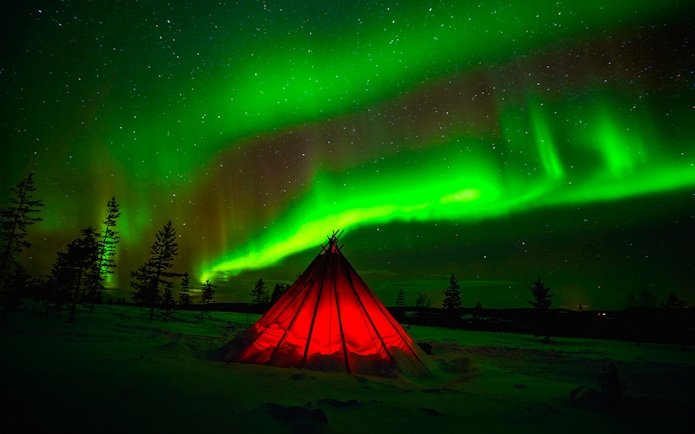 Northern Lights over snowy Rovaniemi landscape with illuminated tent during guided tour.