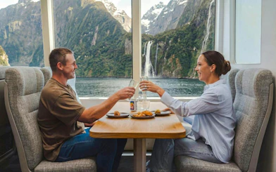 Couple enjoying a meal on a luxury cruise with Milford Sound waterfall view.