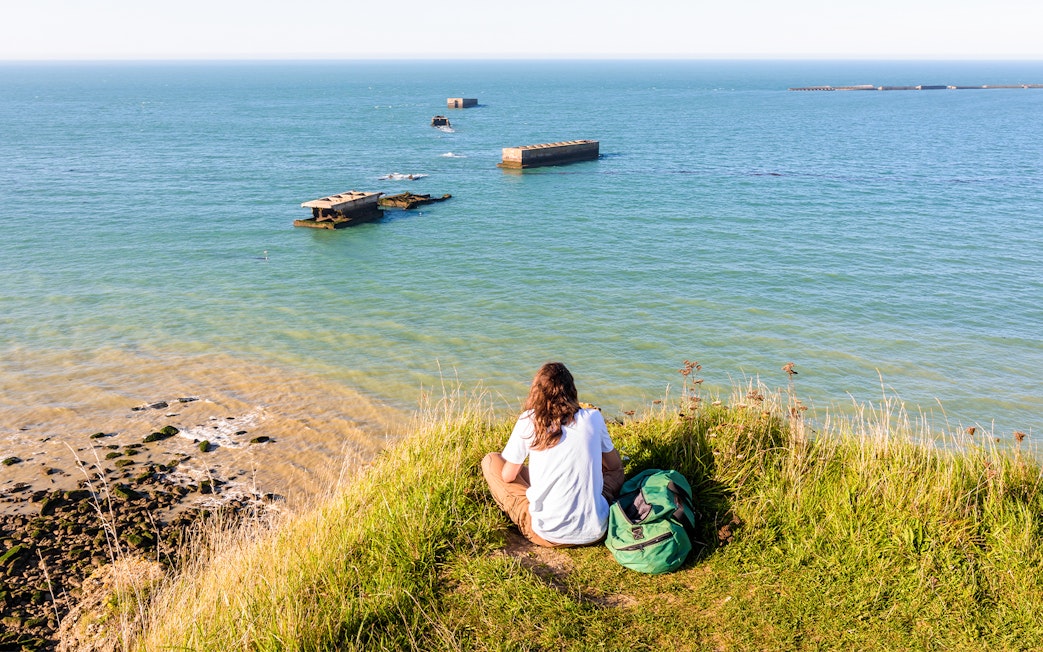Person sitting on a grassy cliff overlooking the Normandy D-Day beaches with WWII remnants in the sea.