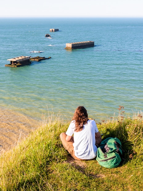 Person sitting on a grassy cliff overlooking the Normandy D-Day beaches with WWII remnants in the sea.