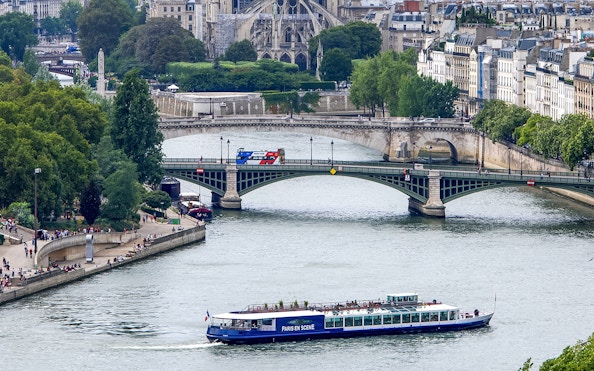 Dinner cruise boat on the Seine River in Paris passing under a bridge with cityscape in the background.