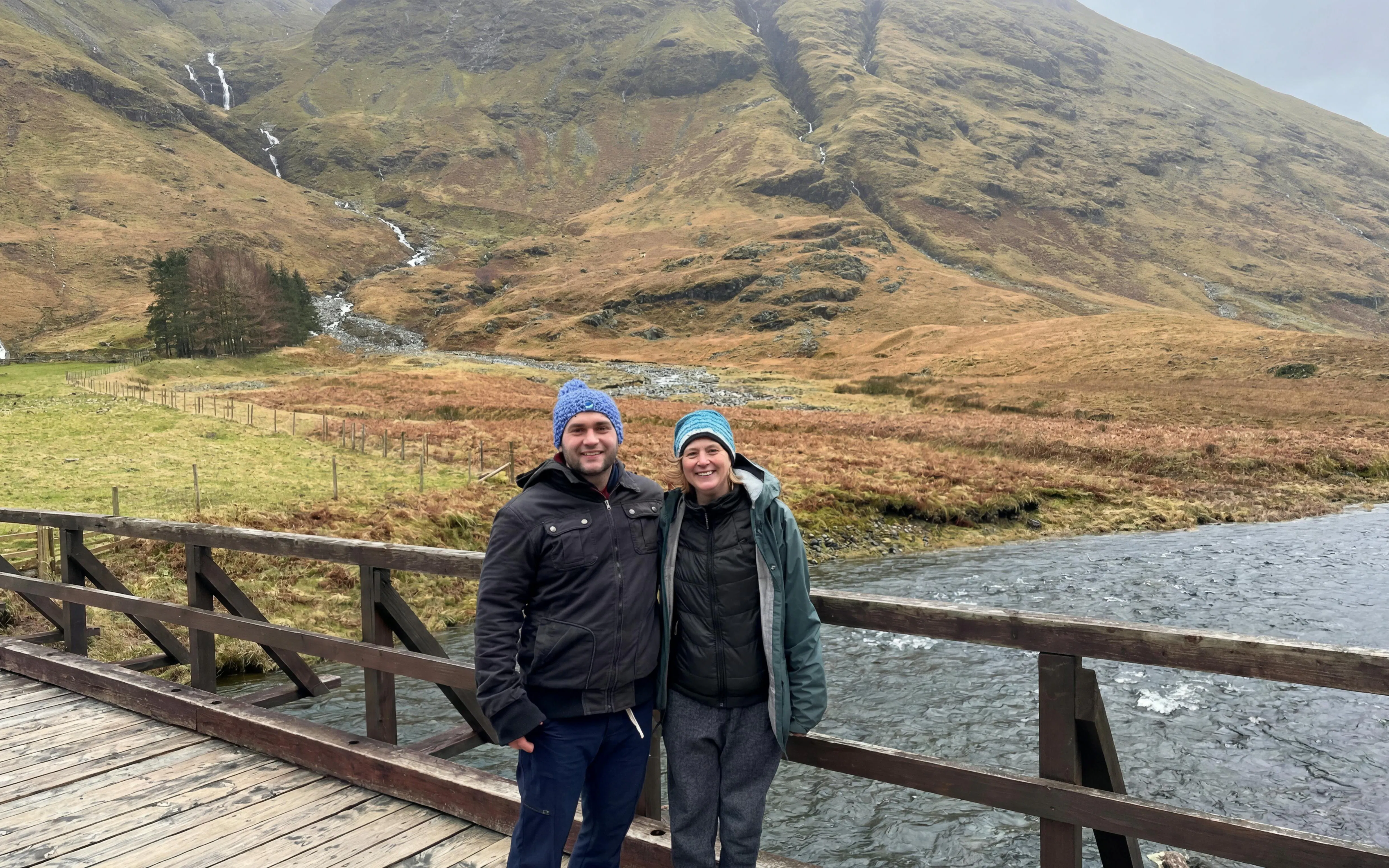 Couple standing on a wooden bridge in Glencoe with mountains in the background.
