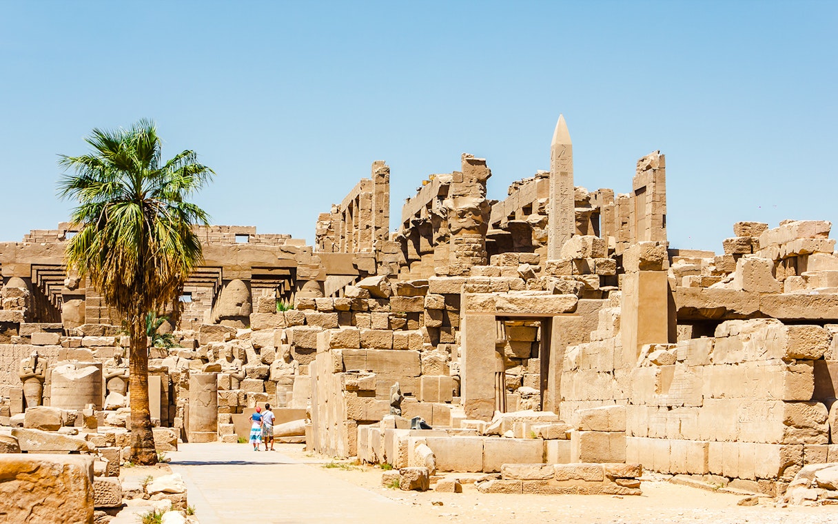 Luxor Temple ruins with palm tree and tourists exploring the ancient site.