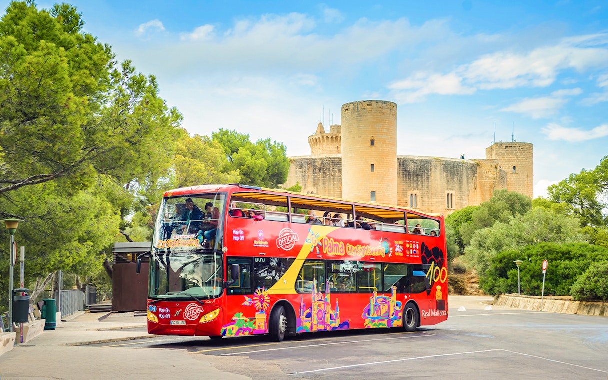 Red double-decker bus on Palma de Mallorca street near Bellver Castle, offering hop-on hop-off city tour.
