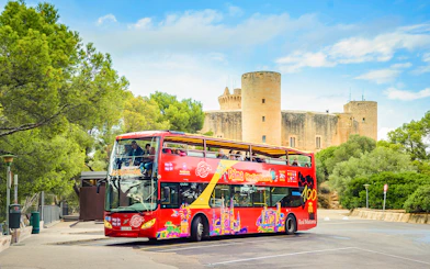 Red double-decker bus on Palma de Mallorca street near Bellver Castle, offering hop-on hop-off city tour.