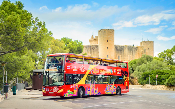 Red double-decker bus on Palma de Mallorca street near Bellver Castle, offering hop-on hop-off city tour.