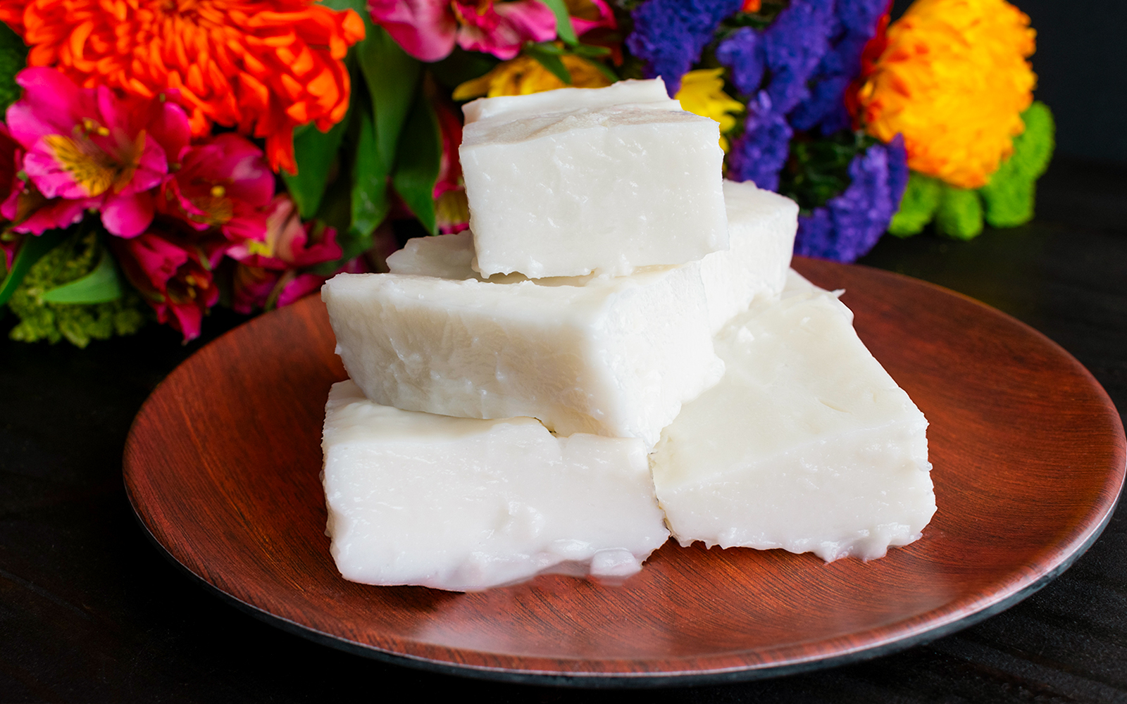 Haupia coconut pudding squares on a wooden plate with colorful flowers in the background.