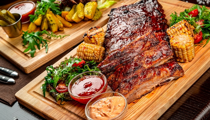 Barbecue ribs with grilled corn, salad, and sauces on a wooden platter.