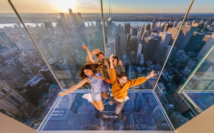 Four people posing on a glass platform above a city skyline at sunset.