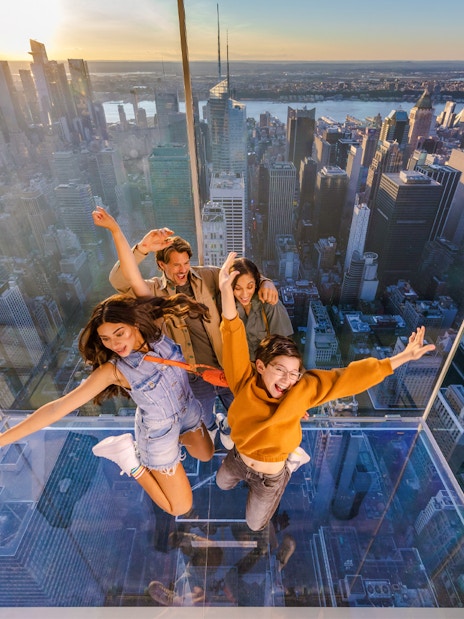 Four people posing on a glass platform above a city skyline at sunset.