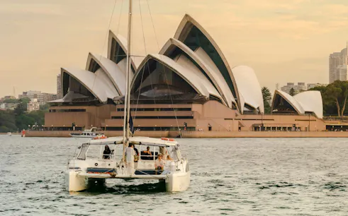 Catamaran sailing near Sydney Opera House at sunset.