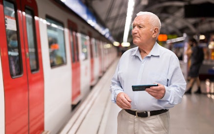 Elderly man holding a phone at a train station platform.